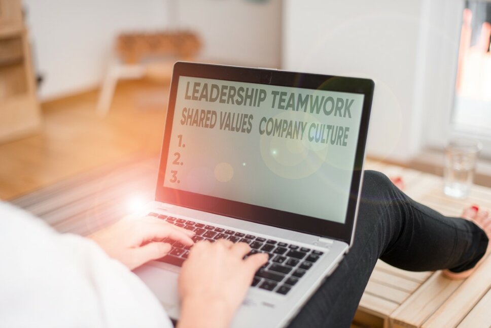 Eine Frau sitzt auf dem Boden mit einem Laptop auf ihrem Schoß. Auf dem Bildschirm sind die Wörter 'Leadership, Teamwork, Shared Values und Company Culture' zu lesen, während das Licht auf dem Laptop reflektiert wird.