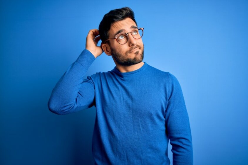 Ein Mann vor einem blauen Hintergrund greift sich nachdenklich an den Kopf und blickt in die Luft, symbolisch für häufig gestellte Fragen (FAQs) im Bewerbungsprozess.