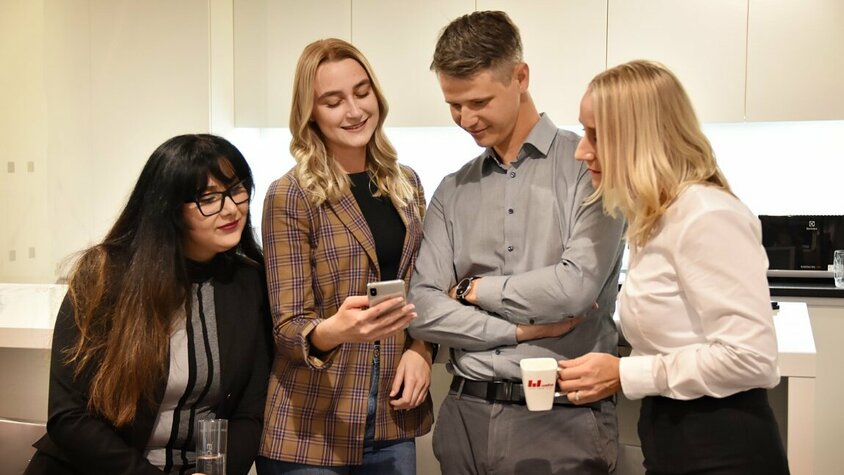 Drei Mitarbeiterinnen und ein Mitarbeiter von Weba Olomouc stehen lächelnd in einer Büroküche und blicken gemeinsam auf ein Handy. Zwei Personen halten eine Kaffeetasse in der Hand, was auf eine entspannte, kollegiale Atmosphäre hindeutet.