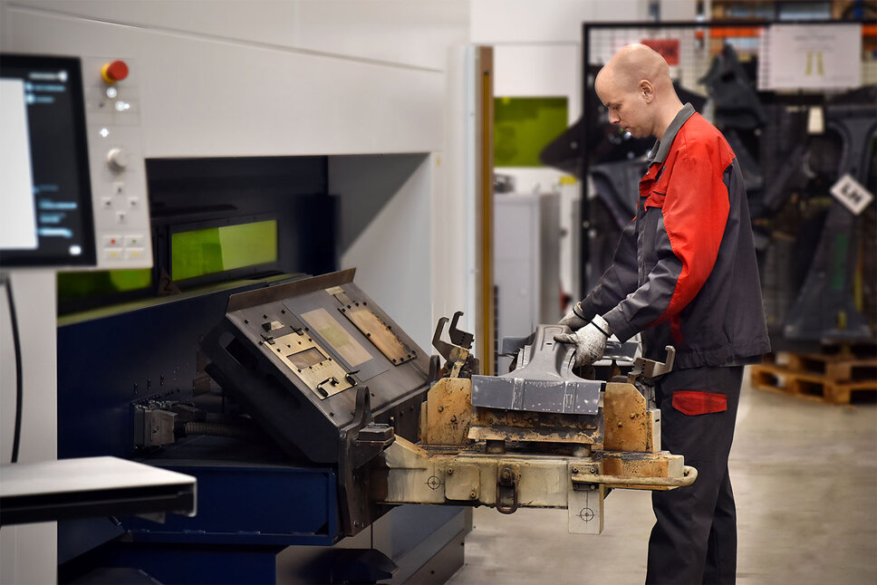 An employee is operating a laser cutting machine in a production hall. He is holding a metal workpiece that is being inserted into the machine for laser cutting. The work environment is clean and well-organized, with various tools and materials in the background.