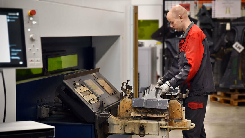 Ein Mitarbeiter bedient eine Laserschneidmaschine in einer Produktionshalle. Er hält ein Werkstück aus Metall, das gerade in die Maschine zum Laserschneiden eingesetzt wird. Die Arbeitsumgebung ist sauber und gut organisiert, mit verschiedenen Werkzeugen und Materialien im Hintergrund.