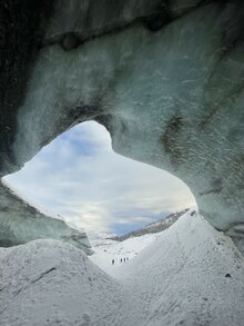 Blick aus einer Gletscherhöhle auf eine verschneite Landschaft mit fernen Bergen. Drei Personen sind in der Ferne sichtbar.