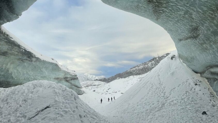 Blick aus einer Gletscherhöhle auf eine verschneite Landschaft mit fernen Bergen. Drei Personen sind in der Ferne sichtbar.