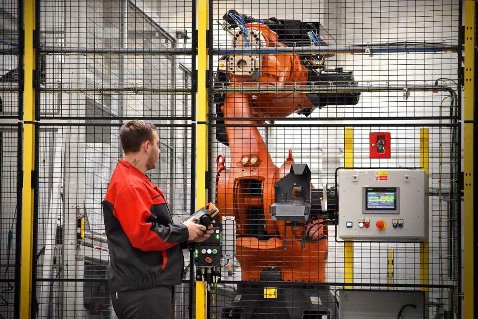 An employee in red workwear is operating an industrial robot in a secured production cell. The robot arm is visible behind a safety fence, and the operator is holding a control device to monitor or adjust the robot's movements. Next to the robot, there is a control panel with a digital display used for managing and monitoring the robotic processes. The scene illustrates the use of automation and advanced robotics in mechanical machining.