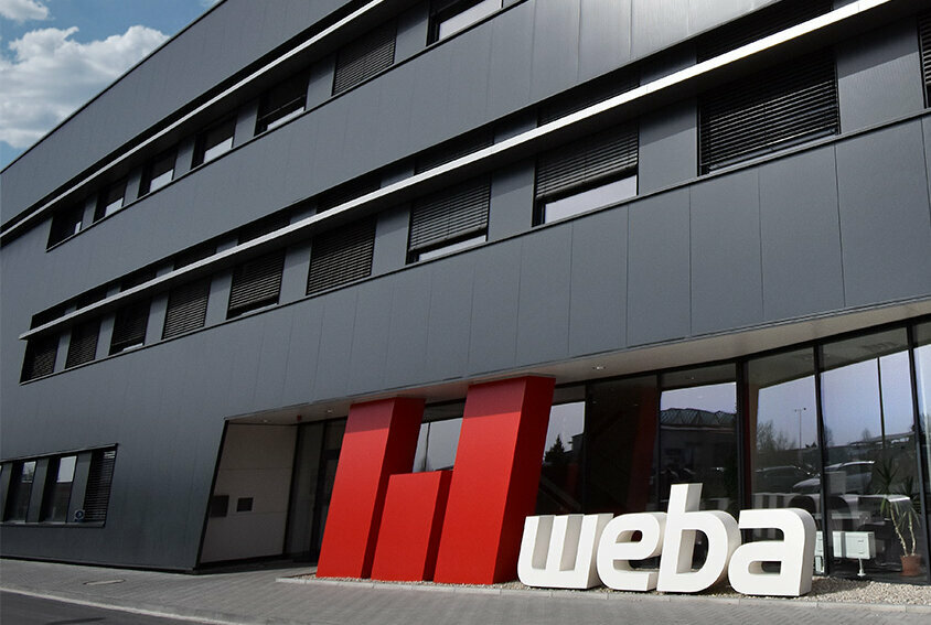Foto der Front des weba-Gebäudes in Olomouc, gekennzeichnet durch eine anthrazitfarbene Fassade. Im Vordergrund steht ein auffälliges 3D-Logo in Rot und Weiß, das die Markenidentität von weba stark hervorhebt und einen markanten Kontrast zur dunklen Gebäudefarbe bildet.