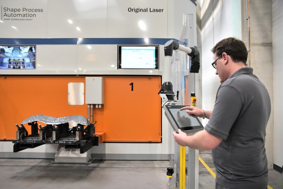 Ein Lasertechniker programmiert die im Hintergrund zu sehende Laserschneidanlage auf einem Tablet