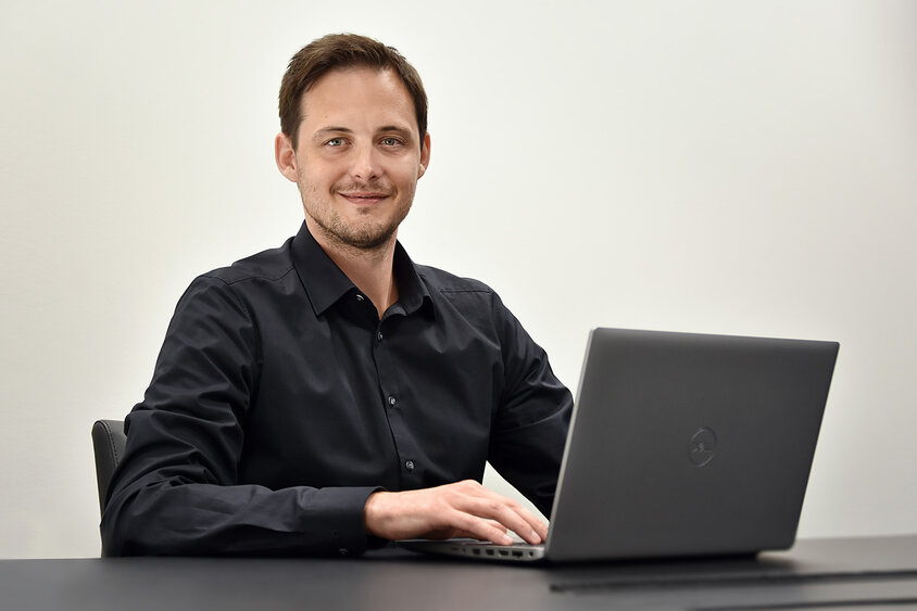 Der HR Manager von weba Olomouc sitzt an seinem Arbeitsplatz, vor ihm ein geöffneter Laptop. Er lächelt freundlich in die Kamera, was eine einladende und professionelle Arbeitsatmosphäre vermittelt. 