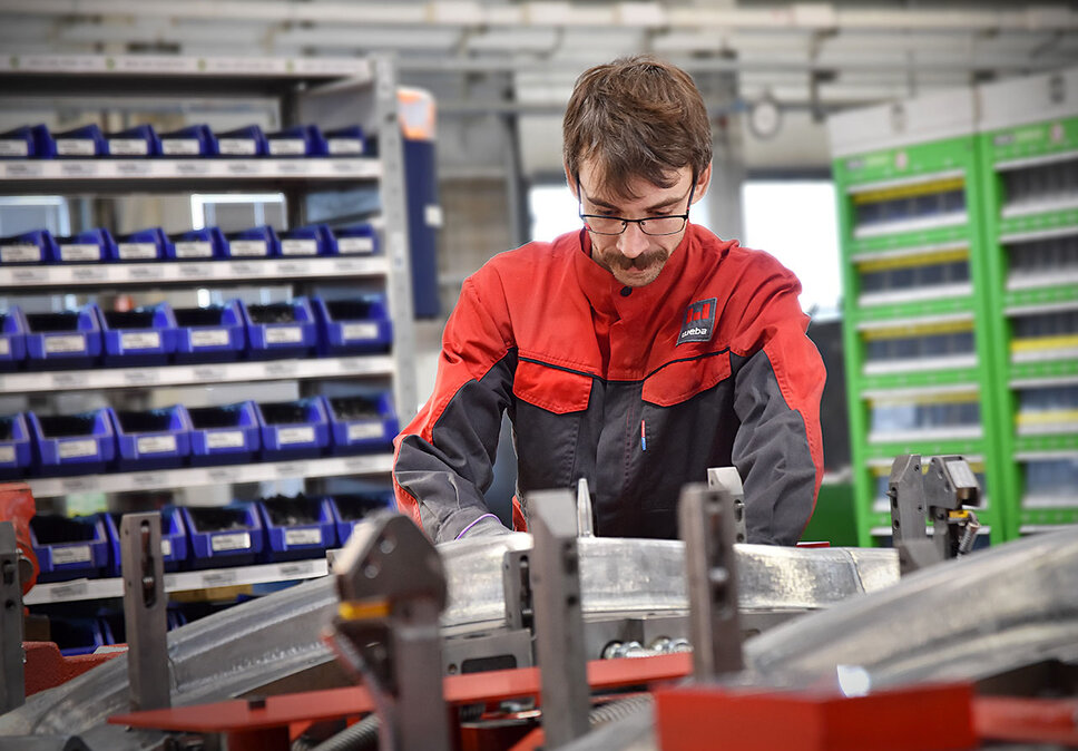  A toolmaking technician is intently assembling a pressing tool for subsequent tool tryout and the forming of structural components. In the background, shelves with auxiliary and operating materials are out of focus.