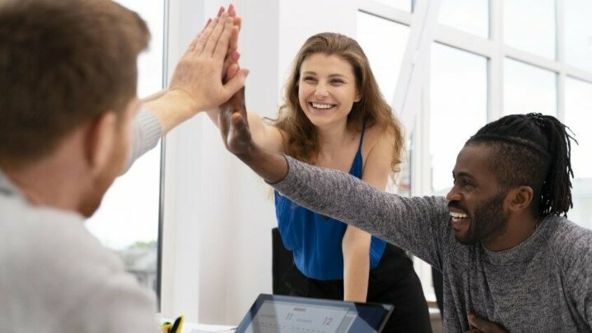 Drei Personen sitzen lächelnd an einem Besprechungstisch und geben sich High-Five.