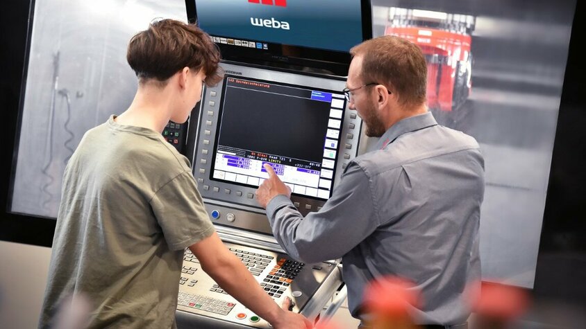 Ein Ausbilder erklärt einem Lehrling in der Zerspanungstechnik die Bedienfunktionen an der Steuerung eines Bearbeitungszentrums. In der Lehre in Steyr bei weba Werkzeugbau liegt der Fokus auf praxisnaher Ausbildung direkt an Maschinen.