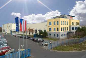 Außenaufnahme des Produktionsstandorts weba Olomouc, bei Sonnenschein unter einem blauen, fast wolkenlosen Himmel, vor dem Gebäude wehen Fahnen.