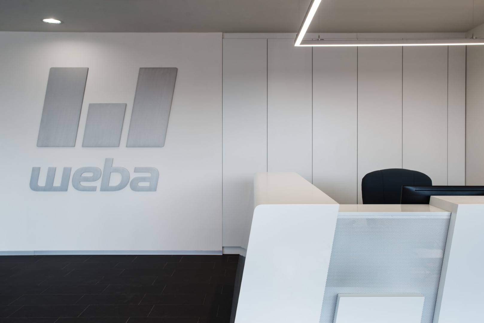 Moderne weiße Empfangstheke im weba-Standort Olomouc, mit dem weba-Logo in gebürstetem Aluminium an der Wand dahinter.