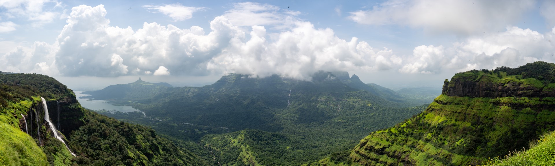 Ein malerisches Panorama mit grünen Hügeln, dichten Wäldern und Wasserfällen, umgeben von einer nebligen Berglandschaft unter einem Himmel mit weißen, dramatischen Wolken.